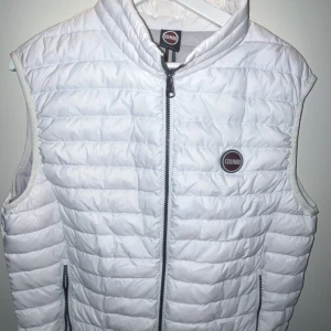 Vit dunväst från Colmar - Snygg vit dunväst från Colmar med quiltad design och dragkedja framtill. Västen har hög krage och två fickor med dragkedja. Perfekt att slänga över en hoodie eller tröja för extra värme. Colmar-logga på bröstet ger en sportig touch.