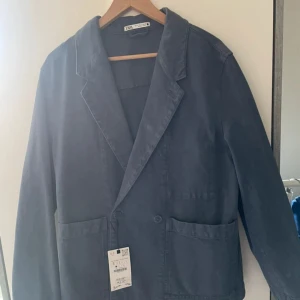 Mörkblå kavaj från Zara, storlek S - Snygg mörkblå kavaj från Zara i storlek S. Klassisk modell med breda slag, två stora framfickor och knappar framtill. Tillverkad i mjuk bomull som ger en avslappnad känsla. Perfekt för dig som gillar stilrena och enkla plagg med modern touch.
