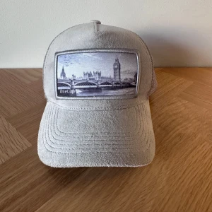 Grå keps från Onecap med Londonmotiv - Snygg grå keps från Onecap med mesh på sidorna och baksidan för extra ventilation. Framtill finns ett broderat motiv av London med Big Ben och en bro, vilket ger en cool urban vibe. Kepsen har böjd skärm och justerbar passform. Perfekt för dig som gillar streetstyle eller en kalssisk stockholm stil.