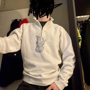 Vit half zip sweatshirt med strassmotiv - Säljer en vit sweatshirt med half zip och lång ärm. På bröstet finns ett detaljerat strassmotiv med en kanin och glittrande stjärnor. Tröjan har en hög krage och är gjord i mjukt material, perfekt för chill dagar. Snygg och unik design som sticker ut.