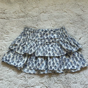 Blåvit mönstrad volangkjol från Zara - Supersöt kort kjol från Zara i storlek 152 men passar xs, med blått mönster på vit botten och dubbla volanger. Kjolen har en smockad midja som gör den bekväm och lätt att bära. Perfekt för dig som vill ha något luftigt och snyggt till sommaren. Bra skick