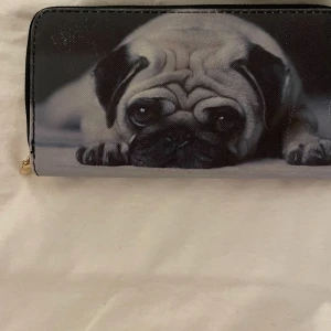 Plånbok med mopsmotiv och dragkedja - Plånbok med ett gulligt mopsmotiv på framsidan. Svart och grå färgton, med guldfärgad dragkedja runtom. Insidan har flera fack för kort och en separat ficka med dragkedja för mynt. Materialet är syntet och ytan är slät.