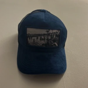 Blå keps med foto-patch - Snygg blå keps med mesh på sidorna och ett svartvitt foto-patch framtill som ger en cool vibe. Perfekt för dig som gillar streetstyle och vill sticka ut med en unik detalj. Kepsen har böjd skärm och justerbar passform.