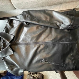 Svart skinnjacka med dragkedja - Snygg svart skinnjacka med klassisk krage och raka linjer. Jackan har två fickor med dragkedja framtill och stilrena sömmar. Perfekt för dig som vill ha en clean och tidlös look. Passar till det mesta och ger en cool vibe.