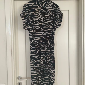 Zebramönstrad långklänning från ONLY - Snygg långklänning från ONLY med svart och beige zebramönster. Klänningen har korta ärmar, knappar framtill och ett matchande bälte i midjan. Perfekt för dig som gillar djurmönster och vill sticka ut. Materialet är mjukt och lätt, vilket gör den skön att bära.