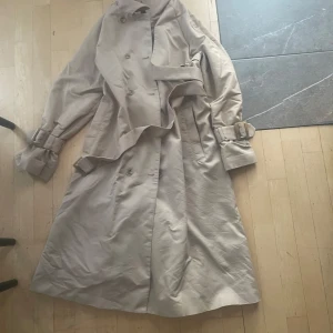 Beige trenchcoat med bälte - Snygg beige trenchcoat med klassisk dubbelknäppning, bälte i midjan och spännen vid ärmsluten. Jackan har en tidlös look och är perfekt för lager-på-lager-stil under kyliga dagar. Lång modell som ger en clean och stilren vibe.