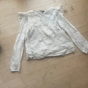 Vit blus med spets och volangdetaljer - Säljer en söt vit blus med långa ärmar, spetsdetaljer över bröstet och volang vid axlarna. Blusen har en romantisk vibe och är gjord i ett lätt bomullstyg som känns luftigt. Perfekt för dig som gillar en feminin och trendig stil.