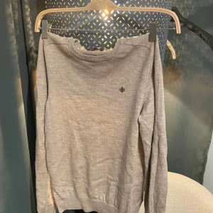 Beige merinoulltröja från Morris - Snygg beige stickad tröja från Morris i 100% merinoull. Klassisk rund hals och diskret brunt broderat emblem på bröstet. Långärmad modell med ribbade muddar och nederkant. Perfekt för dig som gillar stilrena och mjuka plagg.