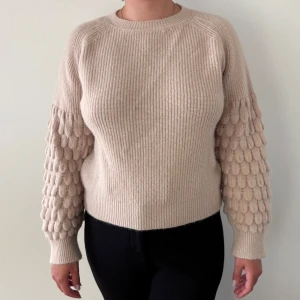 Beige stickad tröja med strukturärmar - Säljer en beige stickad tröja med rund hals och unika ärmar med strukturstickade detaljer som ger en 3D-effekt. Tröjan har ribbade muddar och en loose passform,.