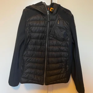 Svart pufferjacka från Parajumpers - Svart cardigan  från Parajumpers med huva och snygga quiltade paneler framtill och baktill. Jackan har dragkedja hela vägen, praktisk bröstficka med zip och detaljer i orange på insidan. Materialmix av polyester och syntet, perfekt för kyliga dagar. Och så har den en bakficka och fickor inuti som är stora ända defekten på jackan är fickorna inne i jackan då sömmen har gått av lite men absolut inget man märker Arnars top skick! Ny pris runt 4000kr. Storlek Y-L har också en parajumper tag med