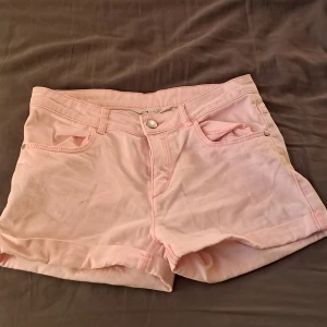 Ljusrosa jeansshorts med fickor - Snygga ljusrosa jeansshorts med klassisk femficksdesign och knappstängning fram. Perfekta för varma dagar och har en skön normal passform. Materialet är mjukt och bekvämt, och färgen ger en fräsch vibe till din outfit.