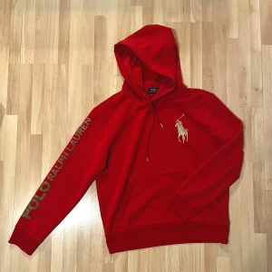 Röd hoodie från Polo Ralph Lauren - Snygg röd hoodie från Polo Ralph Lauren med stort logotryck på ärmen och klassisk polospelare på bröstet. Tröjan har huva med snörning, magficka och ribbade muddar. Perfekt för dig som gillar streetwear och vill sticka ut med en ikonisk look.