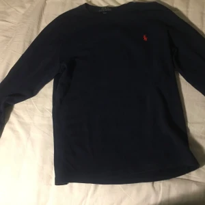 Marinblå långärmad tröja Polo Ralph Lauren - Snygg marinblå långärmad tröja från Polo Ralph Lauren med klassisk röd broderad logga på bröstet. Tröjan är gjord i mjuk bomull och har en enkel, stilren design med rund hals. Perfekt för dig som gillar tidlös och clean stil. Den är L men den är för liten för mig som är 170