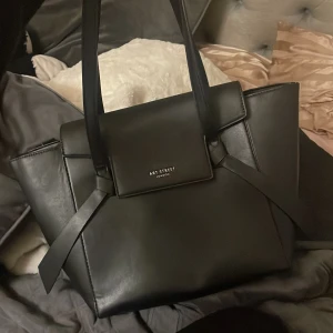 Svart handväska från Art Street London - Snygg svart handväska i skinnimitation från Art Street London. Väskan har stilrena linjer, långa axelband och en cool detalj med knyt framtill. Insidan är fodrad med leopardmönster och har flera fack, inklusive dragkedja på baksidan. Perfekt för dig som gillar trendiga accessoarer.