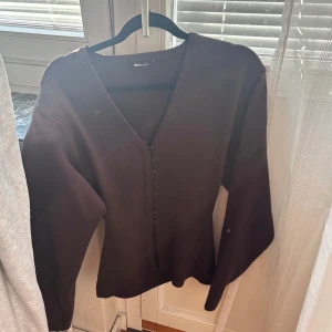 Brun kofta med v-ringning från Gina Tricot - Säljer en mörkbrun kofta från Gina Tricot med v-ringad hals och dekorativa knappar framtill. Koftan har långärmade ärmar och är gjord i ett mjukt stickat material som är perfekt för kyliga dagar. Enkel och stilren design som funkar till många outfits.