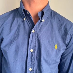Blå skjorta från Polo Ralph Lauren - Snygg blå skjorta från Polo Ralph Lauren i slim fit med klassisk button-down krage och broderad gul logga på bröstet. Skjortan har långa ärmar och vita knappar framtill. Perfekt för dig som gillar stilrena och tidlösa plagg.