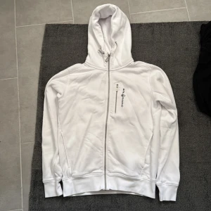 Sail Racing vit hoodie med dragkedja - Vit hoodie från Sail Racing i storlek S. Tröjan har hel dragkedja framtill, en extra ficka med dragkedja på bröstet och broderad logga. Mjuk bomullskänsla, huva med snörning och långa ärmar. Perfekt för chill eller när du vill ha en clean look. Mycket fint skick. 