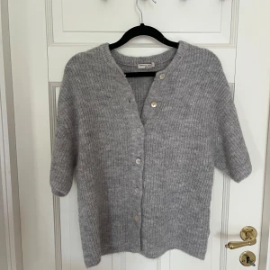 Grå stickad kofta från Gina  - Säljer en chill grå kofta från Gina med korta ärmar. Koftan har rund hals och knäpps med fyra knappar framtill. Perfekt att slänga på sig när det är lite kyligt ute. Materialet är mjukt och stickat för extra comfort.