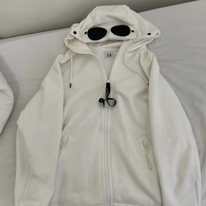 Vit jacka med huva och glasögon från C.P. Company - Säljer en vit kofta från C.P. Company med unik design. Koftan har en huva med inbyggda glasögon och dragkedja framtill. Perfekt för en stilren look med en twist. Köpt i nk i Göteborg använt den 3 gånger bara
