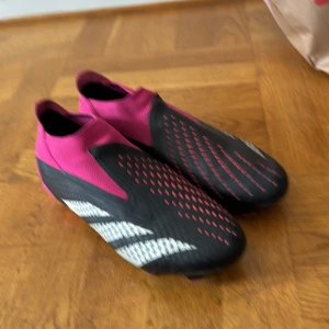 Svart och rosa elit fotbollsskor från Adidas - Snygga fotbollsskor från Adidas i svart och rosa med vita detaljer. Skorna har en strömlinjeformad design utan snörning och en robust sula med dobbar för bra grepp på planen. Perfekta för den som vill sticka ut på fotbollsplanen! Glöm inte det är elit! Har haft skorna i en till två månader och ingen lukt 