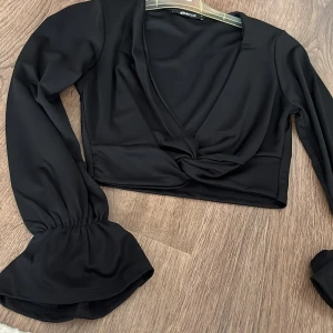 Svart omlottopp från Gina Tricot - Snygg svart omlottopp från Gina Tricot med långa ärmar och v-ringning. Toppen är croppad och har en elegant design som passar perfekt för olika tillfällen.  Kunden står för frakten/ alternativt mötas upp i Norrköping!