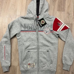 Grå hoodie från Geographical Norway - Säljer en grå hoodie från Geographical Norway med dragkedja och tryckdetaljer. Tröjan har en huva och långa ärmar med ribbade muddar. Det finns röda och vita detaljer på ärmen och bröstet. Perfekt för en avslappnad stil.