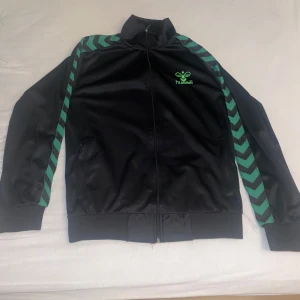 Svart träningsjacka från Hummel - Säljer en svart träningsjacka från Hummel med gröna detaljer. Jackan har en dragkedja framtill och långa ärmar med ett mönster av gröna pilar längs ärmarna. Perfekt för sportiga aktiviteter eller en avslappnad stil.