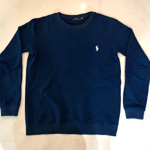 Vintage Blå tröja från Ralph Lauren - Säljer en stilren blå tröja från Ralph Lauren med det klassiska broderade logotypen på bröstet. Tröjan har långa ärmar och ribbade muddar vid ärmslut och nederkant. Perfekt för en avslappnad och snygg look.