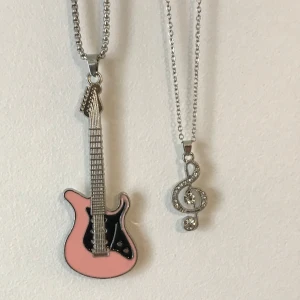 Halsband med gitarr och g-klav - Två unika halsband med musikinspirerade hängen. Det ena har en rosa gitarr med svarta detaljer, och det andra en glittrig g-klav med stenar. Perfekt för musikälskare som vill uttrycka sin stil.