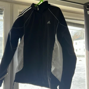 Svart träningsjacka från Adidas - Säljer en svart träningsjacka från Adidas med vita detaljer och dragkedja. Jackan har långa ärmar och är perfekt för sportiga aktiviteter. Den har en klassisk Adidas-design med de ikoniska tre ränderna på ärmarna.