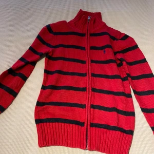 Röd och svart randig tröja från Polo Ralph Lauren - Säljer en snygg röd och svart randig tröja från Polo Ralph Lauren. Tröjan är tillverkad i bomull och har en dragkedja framtill. Perfekt för en stilren look med långa ärmar och ribbade detaljer vid ärmslut och nederkant.