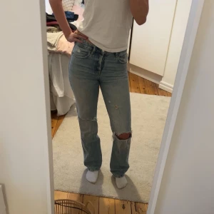 Blå jeans med slitningar - Säljer ett par ljusblå jeans med slitningar på knäna och en avslappnad passform. De har en klassisk femficksdesign och är perfekta för en casual look. Jeansen har en knappgylf och är tillverkade i ett bekvämt denimtyg.