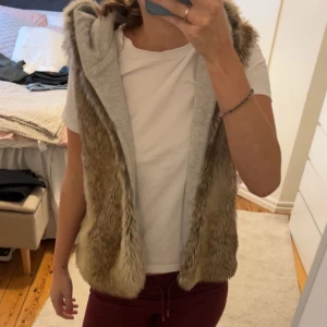 Beige pälsväst med huva - Säljer en mysig beige pälsväst med huva. Västen är ärmlös och har en mjuk insida i grått tyg. Perfekt för att hålla sig varm och stilren under kyligare dagar.