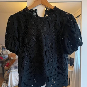 Svart spetsblus - Säljer en elegant svart spetsblus med korta ärmar och ett vackert mönster. Perfekt för att ge en stilfull touch till din outfit. Blusen har en luftig design och är idealisk för varmare dagar.