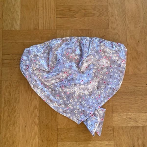 Blommig triangelformad topp  - Snygg blommig lila topp med små vita och rosa blommor med knyte där bak. Tillverkat i 100% polyester och perfekt för att ge en färgglad touch till din outfit. Passar bra för vår och sommar.