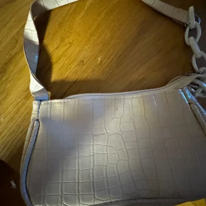 Beige axelväska med krokodilmönster - Snygg beige axelväska med krokodilmönster och dragkedja. Dragkedjan har gått sönder lite.  Väskan har en stilren design med en kedjedetalj på axelremmen. Perfekt för att ge din outfit en trendig touch.