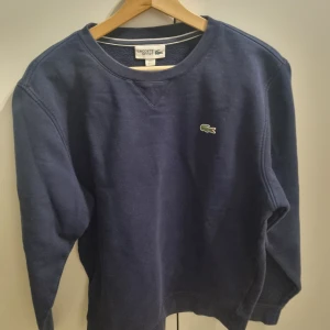 Mörkblå tröja från Lacoste - Mörkblå tröja från Lacoste med det ikoniska krokodilmärket på bröstet. Tröjan har långa ärmar och en rund halsringning.