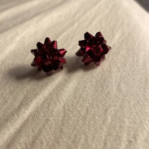 Röda rosettformade örhängen - Snygga örhängen i form av små rosetter i en djup röd färg. Perfekta för att ge en festlig touch till din outfit. De har en enkel stiftfäste för enkel användning.