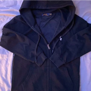 Svart hoodie från Ralph Lauren - Säljer en stilren svart hoodie från Ralph Lauren med dragkedja och det klassiska logotypbroderiet på bröstet. Perfekt för en avslappnad look. Den har en justerbar huva och praktiska fickor framtill. Skicket är bra och inga tecken på defekter eller liknade. Hör av er vid minsta fråga
