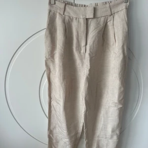 Beige linnebyxor från H&M - Snygga beige linnebyxor från H&M med hög midja och elastisk baksida för extra komfort. Byxorna har en stilren design med veck framtill och diskreta bakfickor. Perfekta för en avslappnad men ändå chic look. Helt oanvända.  Märke: H&M Everyday Färg: Beige Storlek: 40