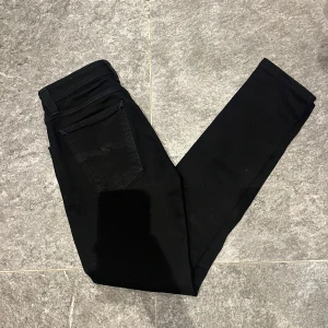 Svarta jeans från Nudie Jeans - Snygga svarta jeans från Nudie Jeans med en klassisk femficksdesign. De har en knappgylf och är tillverkade i ett slitstarkt material. Perfekta för en stilren look. Jeansen är som i nyskick knappt använda pågrund av att dom är för små, modellen är skinny sam och är i st 27/32. Hör av er vid mer frågor.