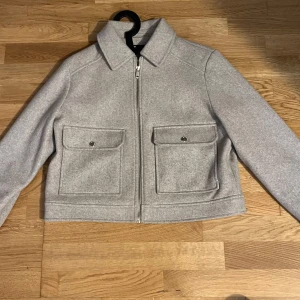 Grå jacka från Zara - Snygg grå jacka från Zara med dragkedja framtill och två stora fickor med knappar. Jackan har en klassisk krage och är perfekt för höst och vår. Den är tillverkad i ett mjukt material som ger en stilren look.