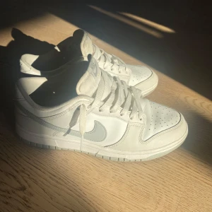 Vita Nike Dunk sneakers - Säljer ett par stilrena vita Nike Dunk sneakers. Skorna har en klassisk design med snörning och en diskret Nike-logga i grått på sidan. Skriv till mig privat om du har frågor.💕