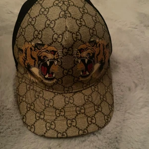 Äkta Gucci keps med tigertryck (pris kan diskuteras) - Säljer en snygg Gucci keps med ett coolt tigertryck. Kepsen har det klassiska GG-mönstret i beige och brunt. Perfekt för att ge din outfit en lyxig touch. Tillverkad i Italien med högkvalitativa material.