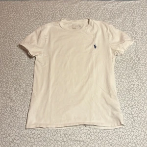 Vit t-shirt från Ralph Lauren - Säljer en klassisk vit t-shirt från Ralph Lauren med den ikoniska broderade loggan i blått på bröstet. T-shirten är kortärmad och tillverkad i mjuk bomull. Perfekt för en stilren och avslappnad look.