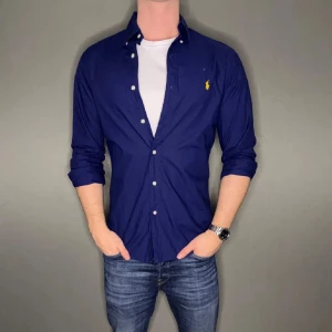 Mörkblå skjorta från Ralph Lauren - Snygg mörkblå skjorta från Ralph Lauren med klassisk krage och knappar framtill. Skjortan har en broderad logga i gult på bröstet och långa ärmar. Perfekt för en stilren look.