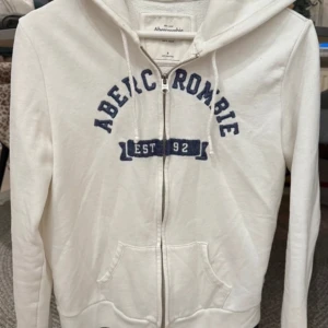 Abercrombie & Fitch kofta - Säljer en vit hoodie från Abercrombie & Fitch med blå text på framsidan. Den har en dragkedja och en klassisk känguruficka. Perfekt för en avslappnad stil.
