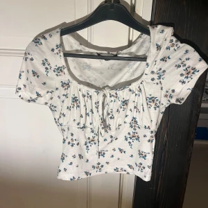 Vit somrig topp - Somrig vit ribbad topp med blommigt mönster i blått. Toppen har korta ärmar och en söt knytning framtill. Står inte storlek men passar nog bra i både S och XS🤍🩵