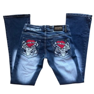 Blå y2k ed hardy liknande jeans med broderade detaljer - Snygga blå jeans med unika broderade dödskallar och hjärtan på bakfickorna med en ripp på ena benet. Midjemått: 36 cm, Längd från gren t midja: 18 cm, Innerbenslängd: 80 cm. Dragkedjan är lite seg, men funkar att dra upp hela vägen om man trycker ihop den lite. Knappen är oxå lite lös men sitter när den väl är knäppt. 