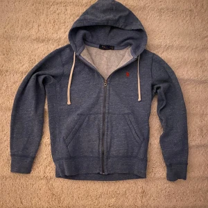 Blå hoodie från Ralph Lauren - Säljer en blå hoodie från Ralph Lauren med dragkedja och justerbar huva. Den har en liten röd logga på bröstet och en mjuk insida. Perfekt för kyliga dagar. 💙notera de lilla hålet på högra armen vid armbågen 